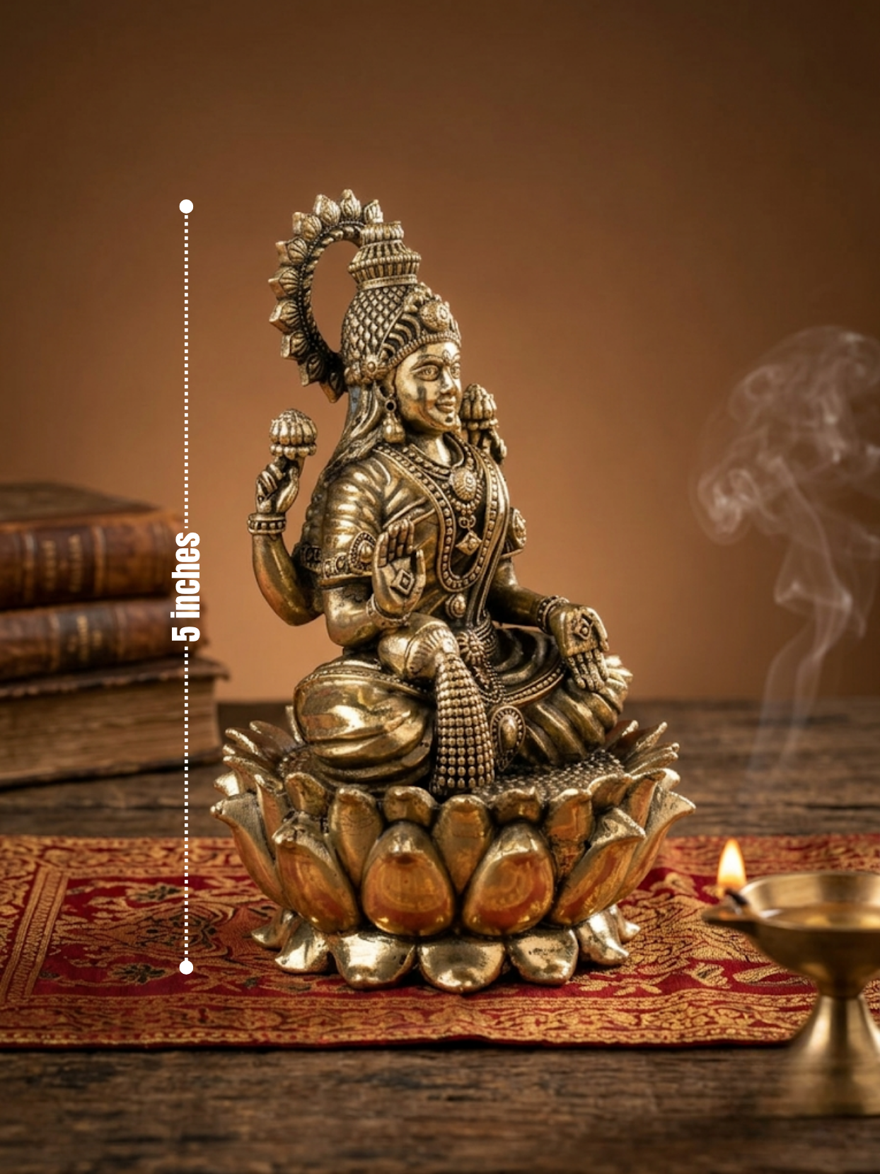 A brass Lakshmi statue seated on a lotus base, displayed on a wooden surface with a red patterned mat, books, and a burning diya in a warm indoor setting. A vertical dotted measurement marker reads 5 inches.