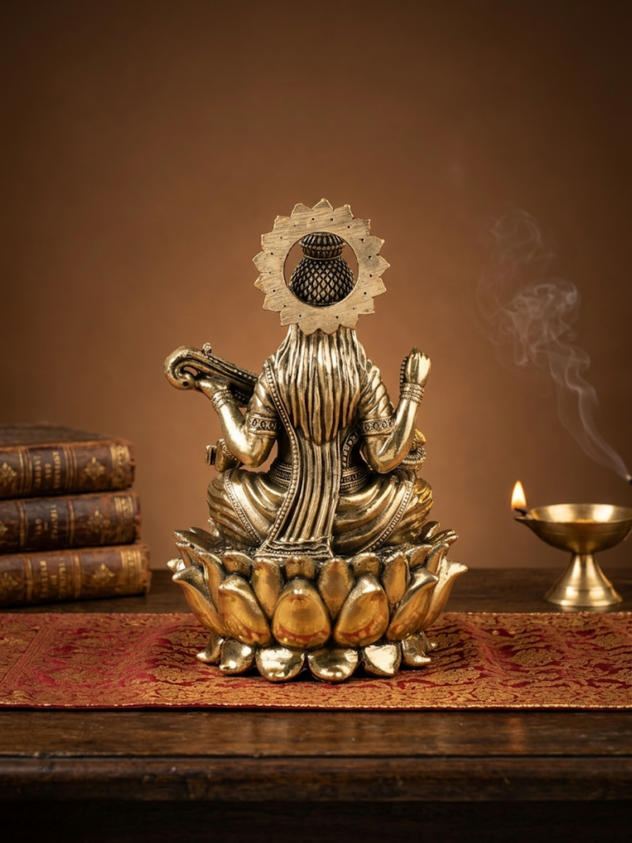 A brass Saraswati idol seated on a lotus base, styled on a wooden table with a red patterned runner, antique books, and a burning diya against a warm brown background.