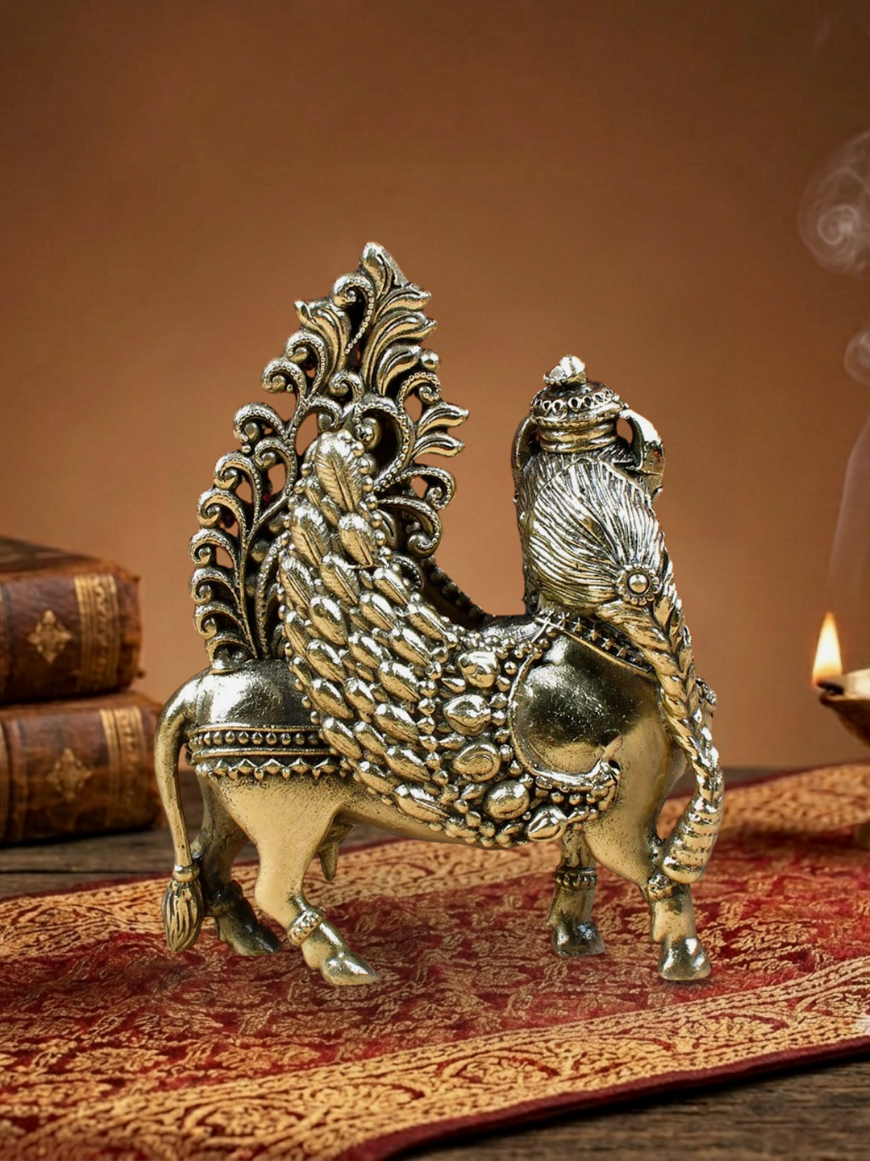 An ornate metallic peacock figurine with a lion-like head, displayed on a red patterned runner with vintage books and a lit oil lamp in the background.