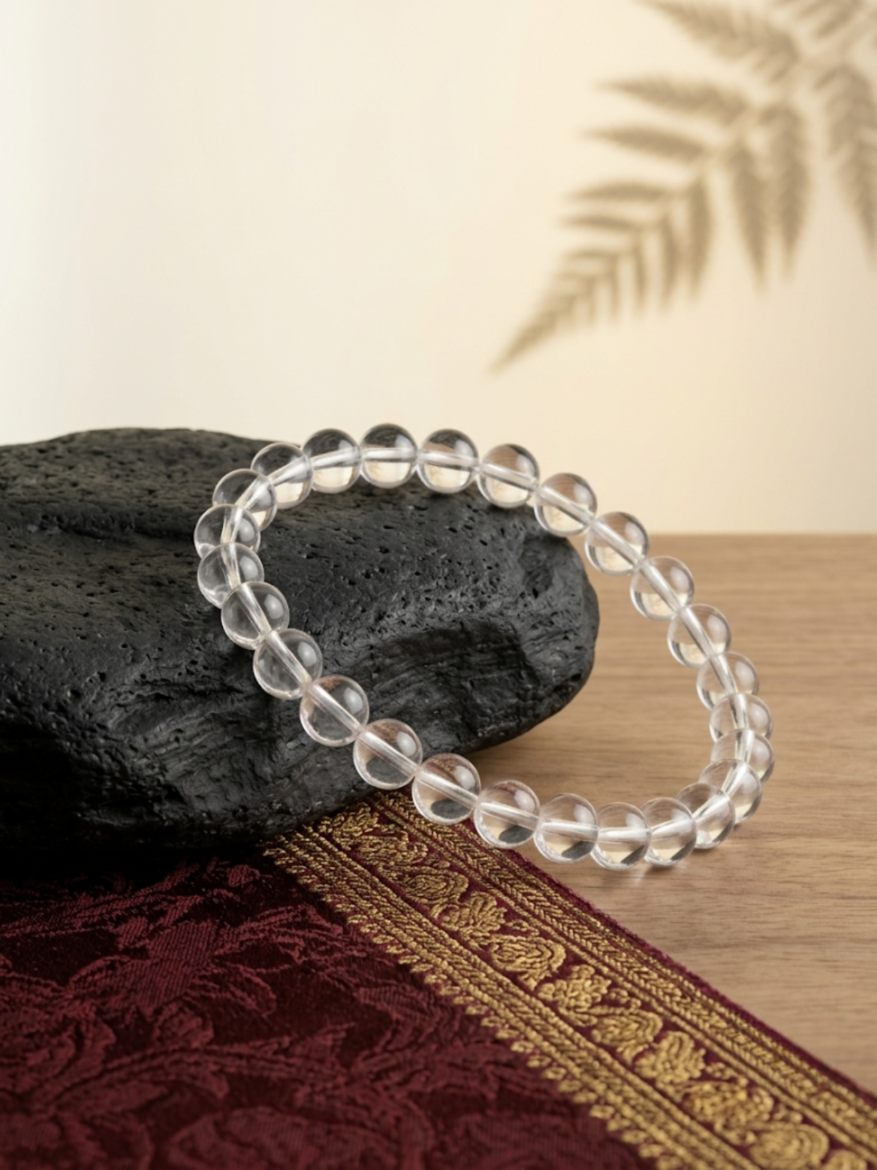 Angled lifestyle shot of the clear 8mm Sphatik beaded stretch bracelet resting on a textured black stone against a warm, neutral background.